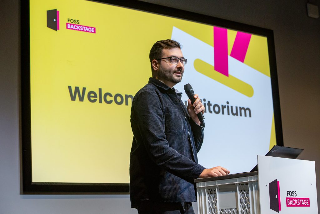 Alexander Brateanu on stage at FOSS Backstage. He is standing at a lectern and holding a microphone.