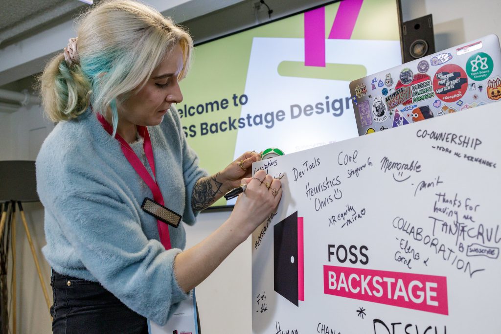 Eriol Fox adding notes to the FOSS Backstage Design lectern sign, which all speakers of the conference have done. On the lectern is a heavily stickered macbook.