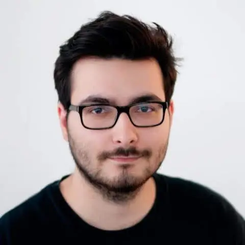 Alex in front of a white background. He has dark hair, a beard, wears glasses and a black tshirt.