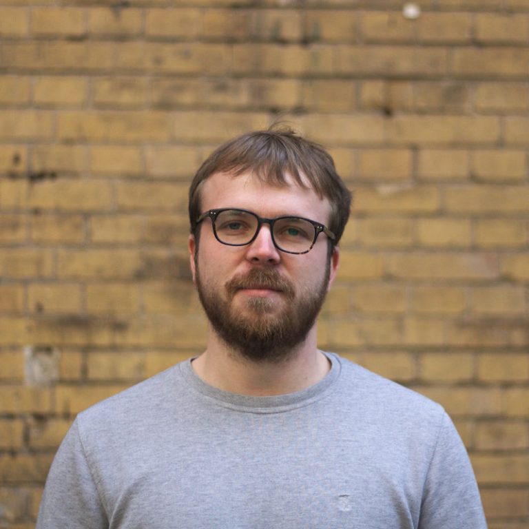 Paul in front of a brick wall. He has a beard, wears a grey sweater and a pair of glasses.